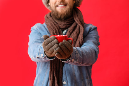 Young bearded man in warm gloves with cup of cocoa on red backgroundの写真素材