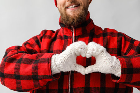 Young man making heart shape with his hands in warm gloves on light backgroundの写真素材