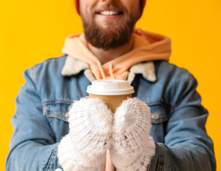 Young bearded man in warm gloves with cup of coffee on yellow background, closeupの写真素材