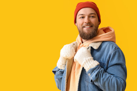 Young bearded man in warm gloves and hat on yellow backgroundの写真素材