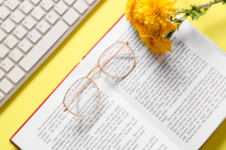 Eyeglasses, computer keyboard, book and flowers on yellow background