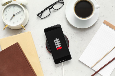 Modern mobile phone charging with wireless pad, cup of coffee and stationery supplies on light...