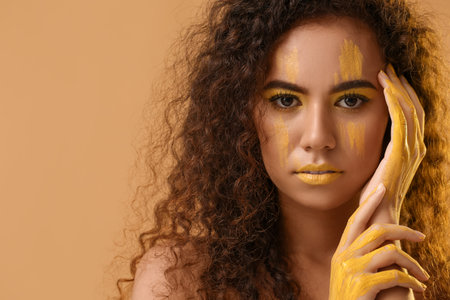 Beautiful young woman with yellow paint on her face and body against color backgroundの写真素材