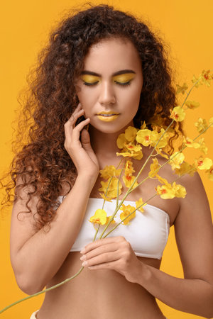 Beautiful African-American woman with makeup in yellow tones and flowers on color backgroundの写真素材