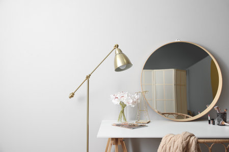 Dressing table with mirror, flowers in vase, cosmetics, jewelry and lamp near light wall