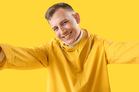 Young man in raincoat taking selfie on yellow backgroundの写真素材