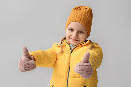 Little girl in stylish winter clothes showing thumb-up gesture on light backgroundの写真素材