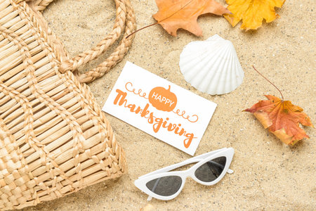 Wicker bag, dry leaves, sunglasses, seashell and paper card with text HAPPY THANKSGIVING on sandの写真素材