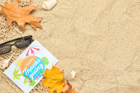 Paper card with text HAPPY THANKSGIVING, nets, sunglasses, dry leaves and seashells on sand backgroundの写真素材