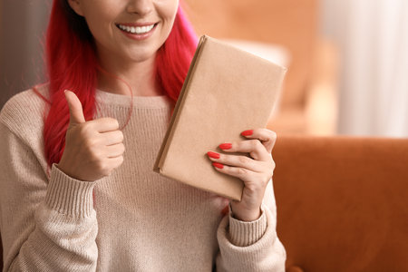 Beautiful woman with book showing thumb-up at home, closeupの写真素材
