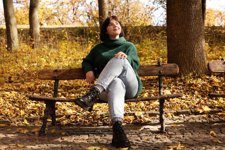 Beautiful woman sitting on wooden bench and enjoying sunny day in autumn parkの写真素材