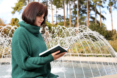 Young beautiful woman reading book near fountain in autumn parkの写真素材