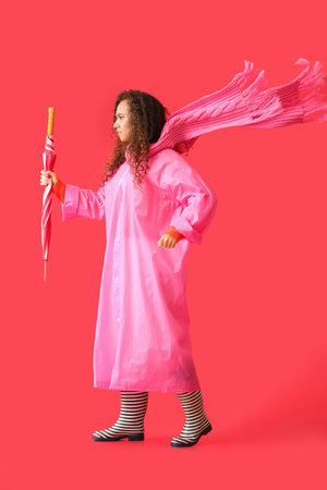 Young African-American woman wearing pink raincoat, scarf and gumboots with umbrella on red backgroundの写真素材