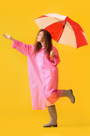 Young African-American woman wearing pink raincoat and gumboots with umbrella on yellow backgroundの写真素材