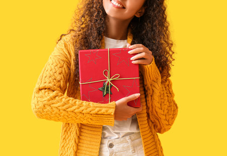 Young African-American woman in warm sweater with Christmas present on yellow backgroundの写真素材