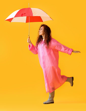 Young African-American woman wearing pink raincoat and gumboots with umbrella on yellow backgroundの写真素材