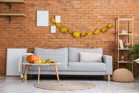 Comfortable sofa, shelf unit and table with pumpkins near brick wall with autumn leaves and blank framesの写真素材