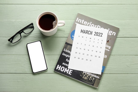 Paper calendar for March 2022, magazine, mobile phone, eyeglasses and cup of coffee on color wooden...