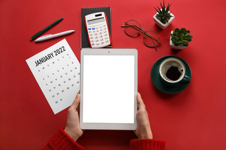 Female hands with tablet computer, paper calendar for January 2022, calculator, eyeglasses and cup of coffee on color backgroundの写真素材