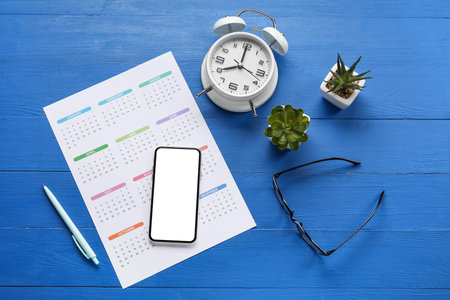 Paper calendar for 2022 year, mobile phone, alarm clock, houseplants, pen and eyeglasses on color wooden backgroundの写真素材