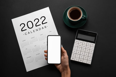 Female hand with mobile phone, paper calendar for 2022 year, cup of coffee and calculator on dark backgroundの写真素材