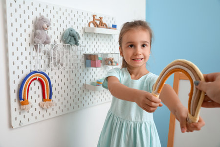 Cute little girl with toy rainbow near hanging peg board on light wallの写真素材