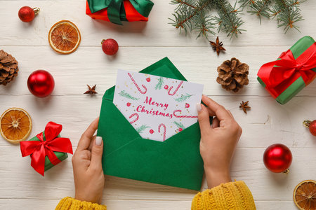 Woman holding envelope and card with text MERRY CHRISTMAS on white wooden table