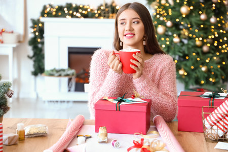 Beautiful young woman wrapping Christmas gift and drinking hot chocolate at homeの写真素材