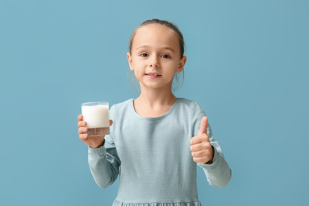 Little girl holding glass of milk and showing thumb up on blue backgroundの写真素材