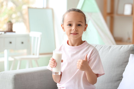 Little girl holding glass of milk and showing thumb up on sofaの写真素材