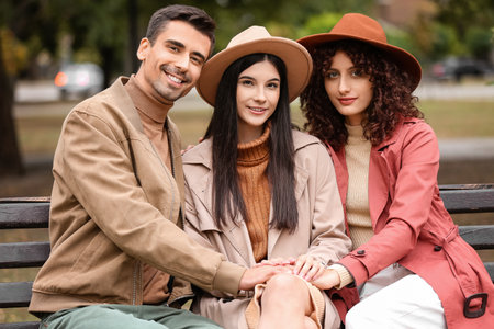 Man and two beautiful women sitting on bench in park. Polyamory conceptの写真素材