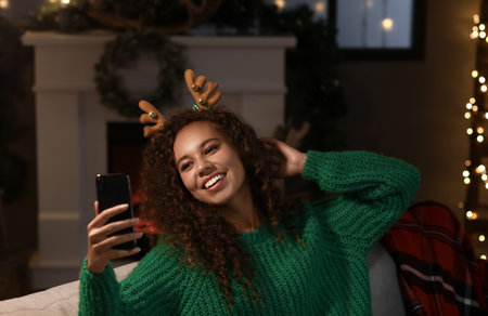 Young African-American woman in deer horns taking selfie at home on Christmas eveの写真素材