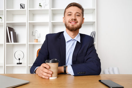 Handsome young businessman with cup of coffee video chatting in office