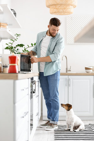 Young man with cute dog adjusting microwave oven in kitchenの写真素材