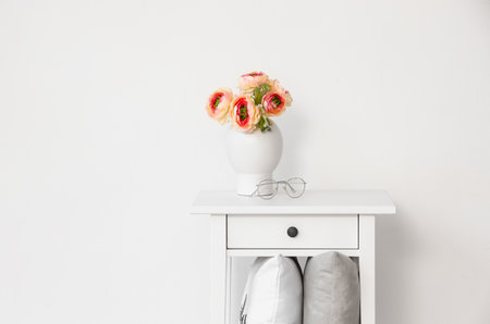 White bedside table with flowers in vase and pillows on shelf near light wall