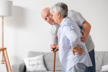 Senior woman with walking stick suffering from back pain and her husband at homeの写真素材