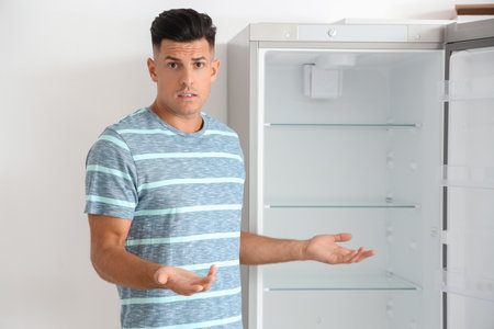 Confused man near empty fridge in kitchenの写真素材