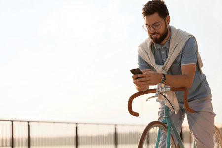 Young bearded man with bicycle using mobile phone on embankmentの写真素材