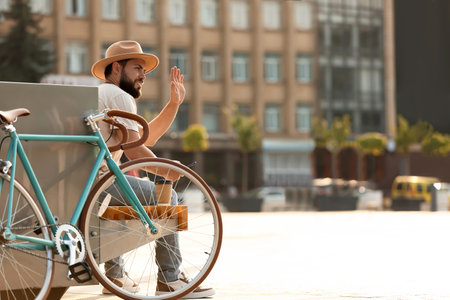 Handsome bearded man with bicycle waving hand on city squareの写真素材