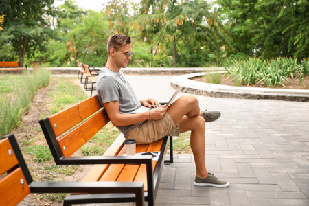 Handsome young man in sunglasses using laptop on bench in parkの写真素材