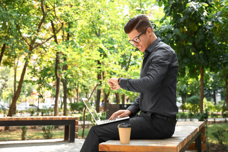 Handsome businessman with laptop looking at wristwatch in parkの写真素材