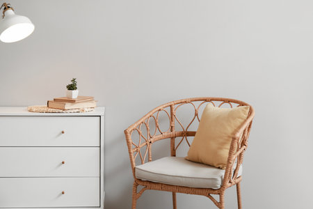 Wicker chair with pillow and chest of drawers near light wall