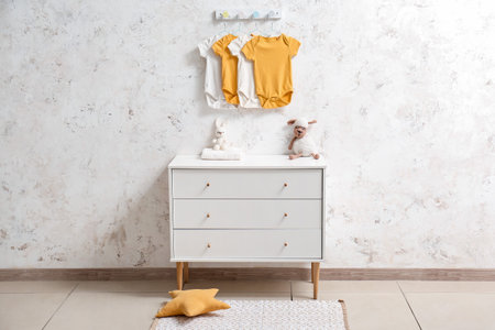 Chest of drawers with toys and baby bodysuits hanging on light wall