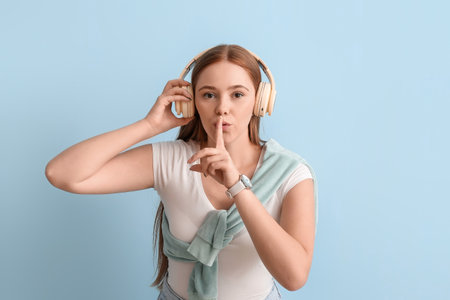 Cool young woman listening to music and showing silence gesture on color background