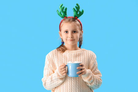 Happy little girl in reindeer horns with cup of cocoa on blue backgroundの写真素材