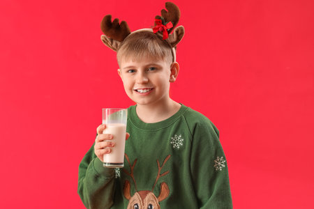 Happy little boy in reindeer horns with glass of milk on red backgroundの写真素材