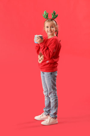 Happy little girl in reindeer horns with cup of cocoa on red backgroundの写真素材