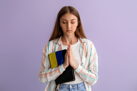 Young woman with Ukrainian flag and Holy Bible praying on lilac backgroundの写真素材