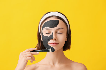 Young woman applying activated charcoal mask on her face against color backgroundの写真素材