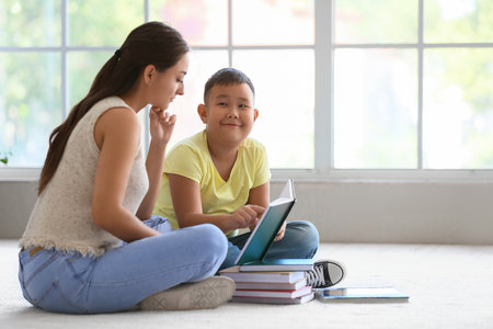 Little Asian boy reading book with his teacher in classroom